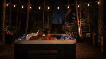 A couple relaxes in a hot tub at night. There are cafe lights overhead and trees in the background.