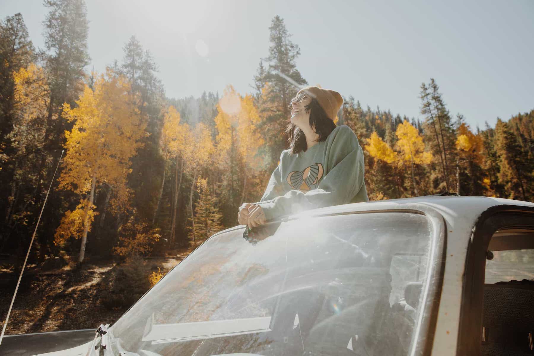A young woman in a cotton beanie standing up through the sunroof of her car. The sun is shining on a beautiful fall day with colorful trees in the background.
