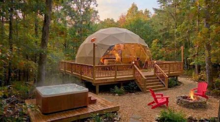An exterior shot of one of Camp Yellow Cardinal's campsites. There is a large wooden deck with a geodesic dome in the center background. Red Adirondack chairs are arranged around the fire-pit. A hot tub sits in the foreground on a low wooden deck.
